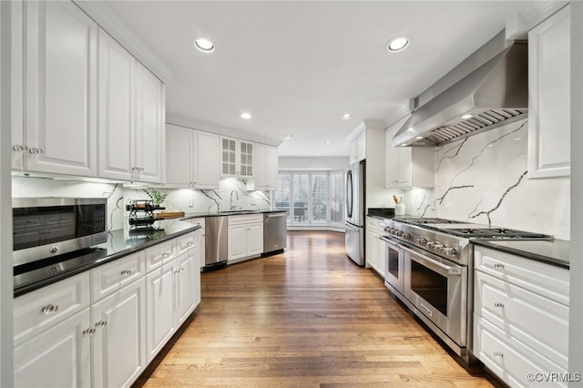 kitchen with stainless steel appliances, white cabinetry, wall chimney exhaust hood, dark countertops, and glass insert cabinets