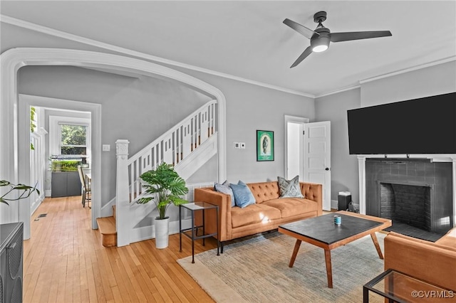 living area with stairs, ornamental molding, a tiled fireplace, light wood-type flooring, and ceiling fan