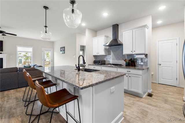 kitchen featuring wall chimney range hood, sink, light stone countertops, an island with sink, and white cabinets