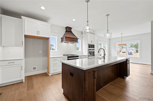 kitchen featuring white cabinetry, decorative light fixtures, an island with sink, custom range hood, and stainless steel appliances