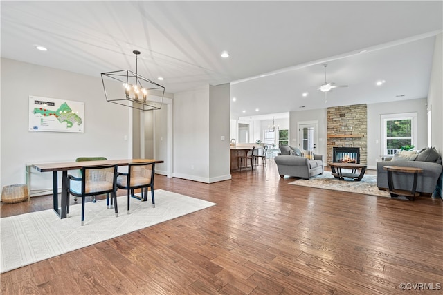 dining area featuring a stone fireplace, ceiling fan with notable chandelier, and dark hardwood / wood-style flooring