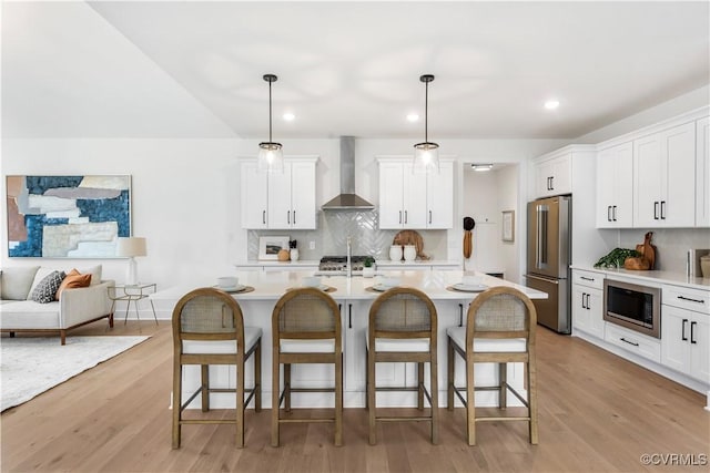 kitchen with white cabinetry, hanging light fixtures, an island with sink, stainless steel appliances, and wall chimney range hood