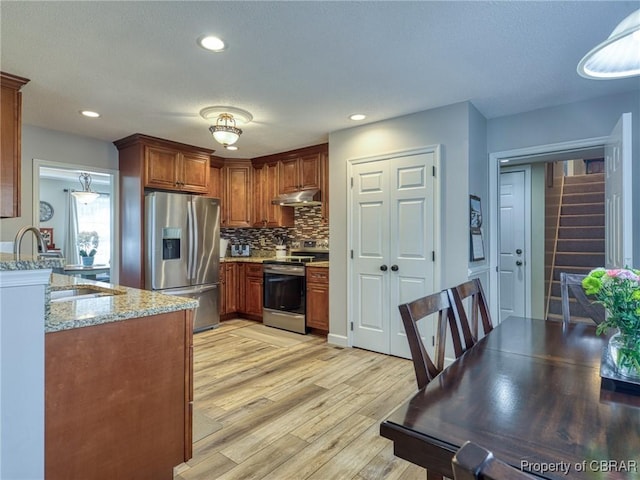 kitchen with appliances with stainless steel finishes, brown cabinets, light stone counters, under cabinet range hood, and pendant lighting