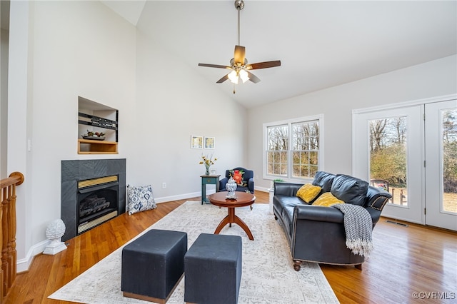 living area with baseboards, ceiling fan, wood finished floors, a fireplace, and high vaulted ceiling