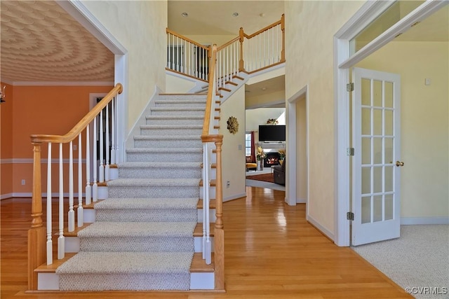 staircase featuring a warm lit fireplace, baseboards, wood finished floors, a high ceiling, and crown molding