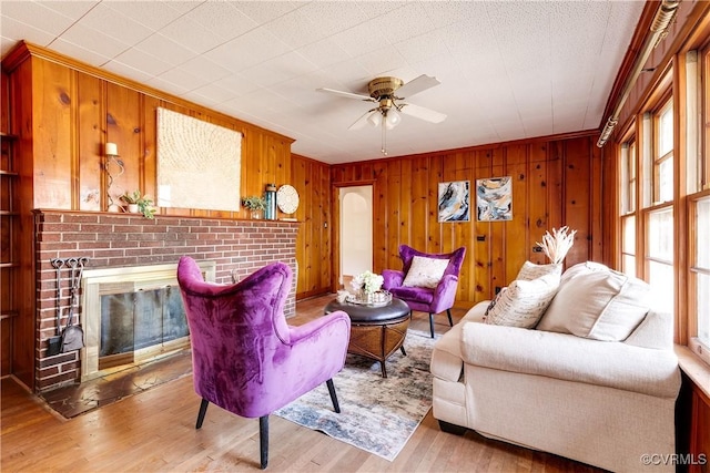 living area with light wood-style floors, a brick fireplace, a ceiling fan, and wood walls