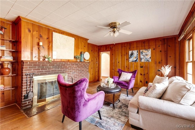 living room with ceiling fan, light wood finished floors, a brick fireplace, and wooden walls