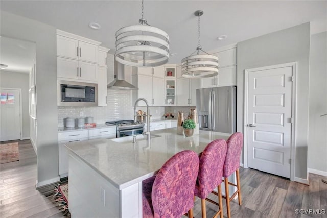 kitchen featuring wall chimney range hood, stainless steel appliances, white cabinets, and a center island with sink