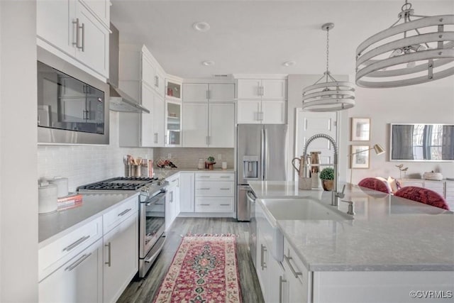 kitchen featuring wall chimney exhaust hood, white cabinetry, decorative light fixtures, stainless steel appliances, and a kitchen island with sink