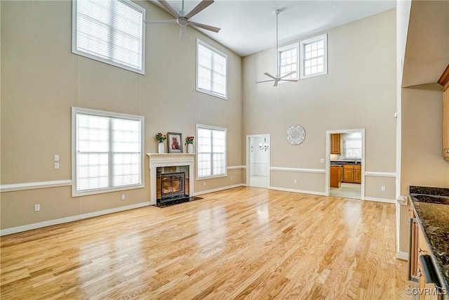 unfurnished living room with a towering ceiling, a ceiling fan, a high end fireplace, light wood-type flooring, and baseboards