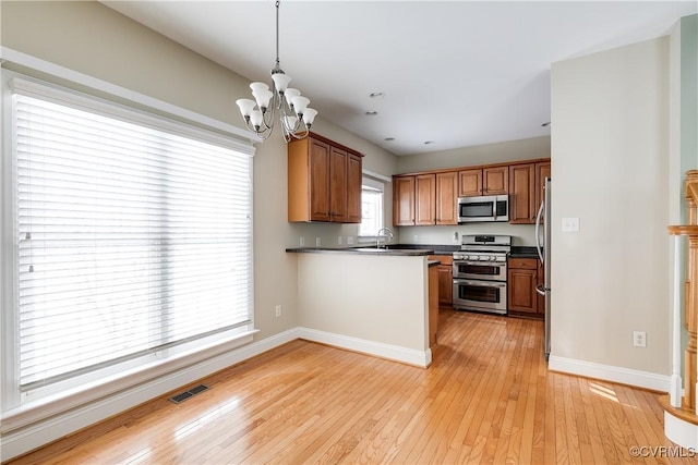 kitchen featuring a peninsula, appliances with stainless steel finishes, brown cabinetry, dark countertops, and decorative light fixtures