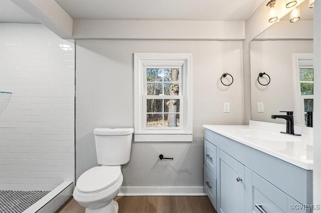 bathroom with toilet, wood finished floors, vanity, baseboards, and tiled shower