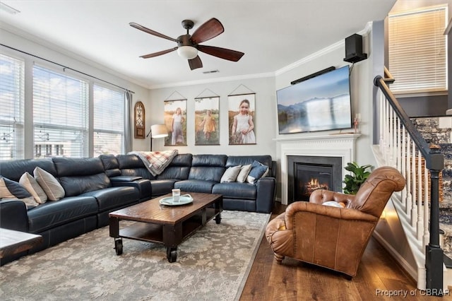 living room with stairway, ornamental molding, a glass covered fireplace, a ceiling fan, and wood finished floors