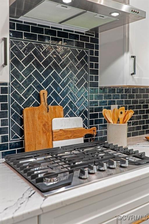 details featuring light stone counters, range hood, stainless steel gas stovetop, backsplash, and white cabinetry