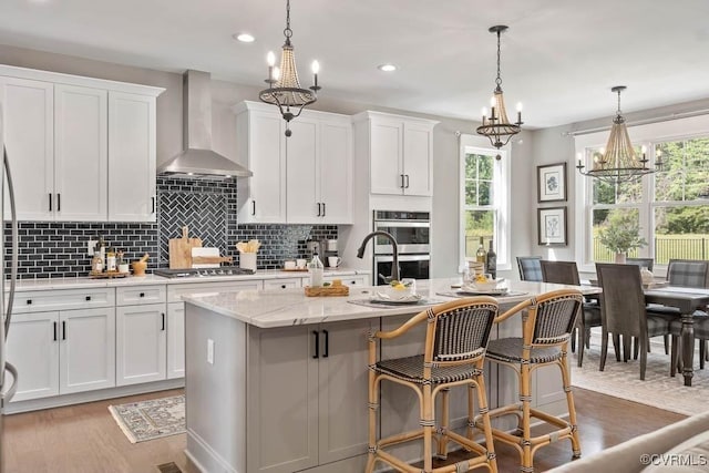 kitchen with a kitchen island with sink, white cabinets, wall chimney range hood, appliances with stainless steel finishes, and backsplash