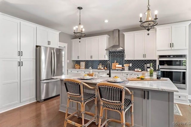 kitchen featuring a kitchen island with sink, stainless steel appliances, wood finished floors, wall chimney range hood, and backsplash