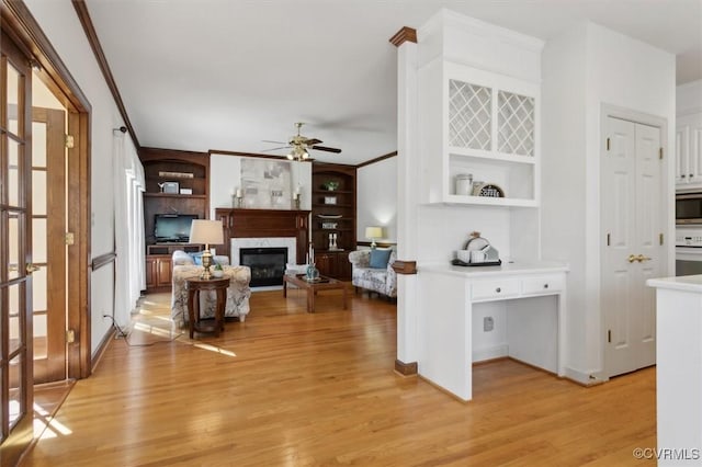 living room with built in shelves, light wood-style flooring, a premium fireplace, ornamental molding, and a ceiling fan