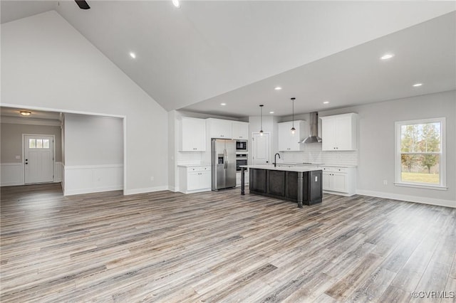 kitchen featuring a center island with sink, stainless steel appliances, white cabinets, wall chimney range hood, and pendant lighting