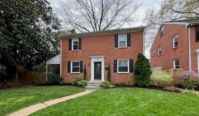 colonial inspired home featuring brick siding, a chimney, a front yard, and fence