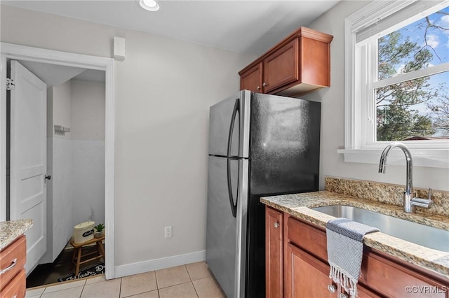 kitchen featuring light stone counters, brown cabinetry, freestanding refrigerator, light tile patterned flooring, and a sink