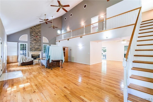 unfurnished living room featuring stairway, a ceiling fan, a stone fireplace, light wood-type flooring, and a towering ceiling