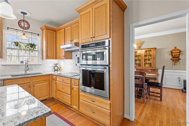 kitchen with light wood finished floors, light stone counters, stainless steel double oven, under cabinet range hood, and a sink