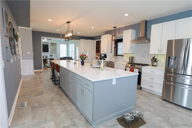 kitchen with a kitchen island with sink, white cabinets, light countertops, appliances with stainless steel finishes, and wall chimney range hood