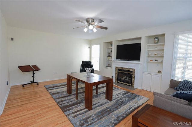 living area with built in shelves, a fireplace with flush hearth, ceiling fan, wood finished floors, and baseboards