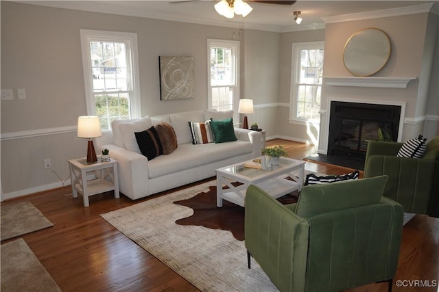 living room featuring wainscoting, dark wood-style floors, a fireplace with flush hearth, ceiling fan, and ornamental molding