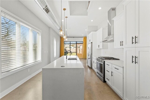 kitchen with pendant lighting, stainless steel appliances, light countertops, a kitchen island with sink, and wall chimney range hood