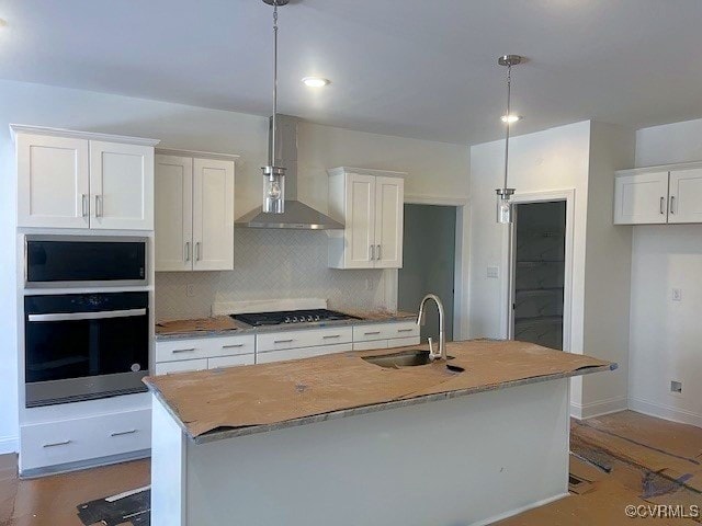 kitchen featuring white cabinets, hanging light fixtures, appliances with stainless steel finishes, wall chimney exhaust hood, and an island with sink