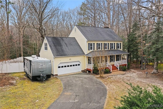 colonial-style house featuring driveway, fence, covered porch, an attached garage, and a shingled roof