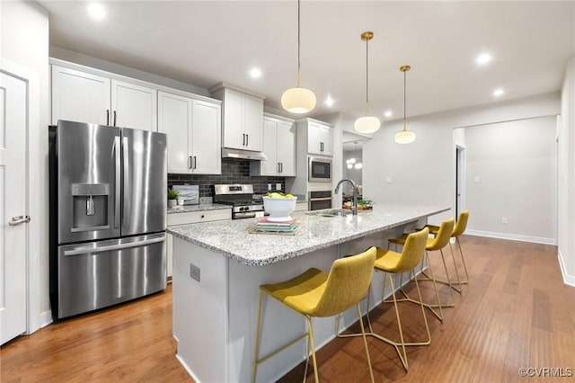 kitchen with a center island with sink, under cabinet range hood, a kitchen breakfast bar, tasteful backsplash, and appliances with stainless steel finishes