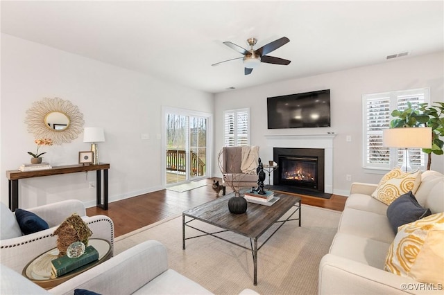 living area featuring baseboards, a fireplace with flush hearth, wood finished floors, and a ceiling fan