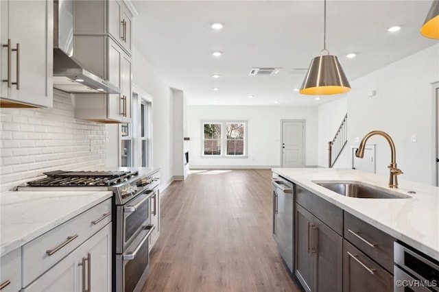 kitchen featuring light stone counters, decorative light fixtures, appliances with stainless steel finishes, a sink, and wall chimney range hood