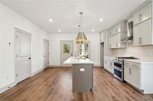 kitchen with light countertops, double oven range, wall chimney exhaust hood, an island with sink, and decorative light fixtures