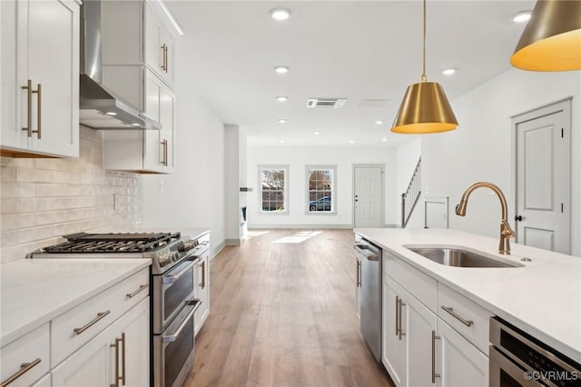 kitchen with hanging light fixtures, appliances with stainless steel finishes, white cabinets, a sink, and wall chimney range hood