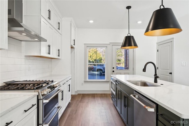 kitchen with appliances with stainless steel finishes, white cabinets, hanging light fixtures, and wall chimney range hood