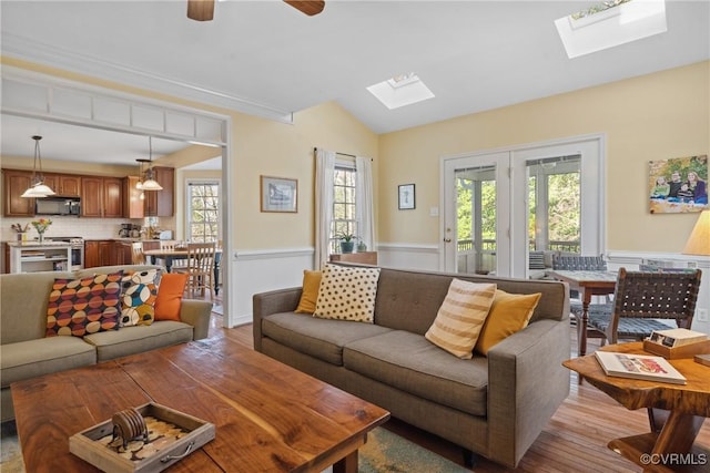 living room featuring vaulted ceiling with skylight, a ceiling fan, wood finished floors, and wainscoting