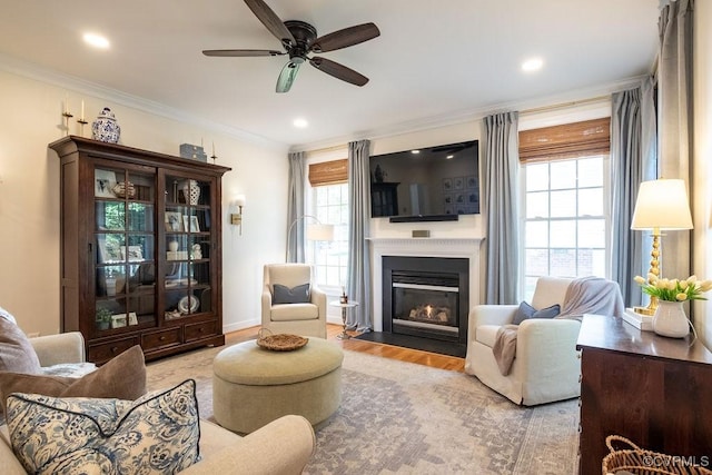 living area with ceiling fan, wood finished floors, a fireplace with flush hearth, baseboards, and ornamental molding