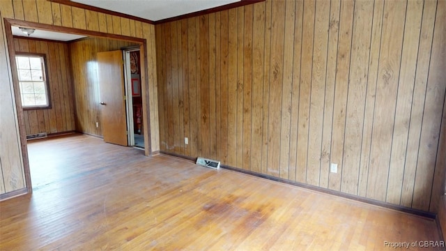 empty room featuring crown molding, light wood finished floors, visible vents, wooden walls, and baseboards