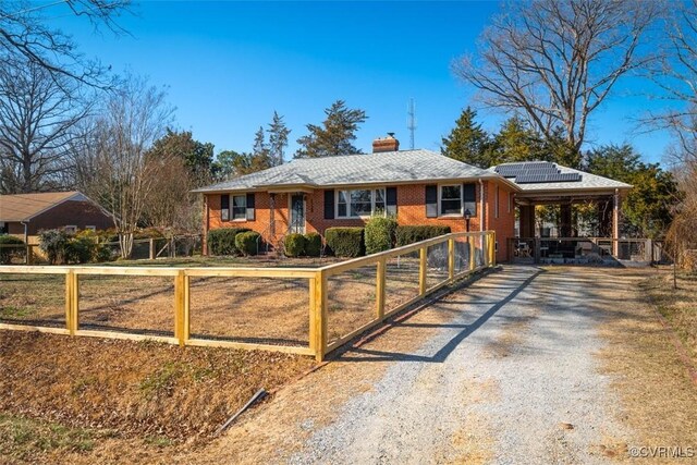 ranch-style house featuring brick siding, gravel driveway, fence, roof mounted solar panels, and a chimney