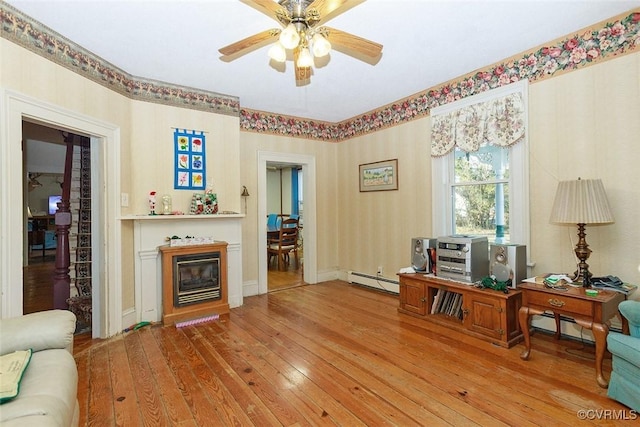 sitting room featuring light wood-type flooring, a baseboard heating unit, a ceiling fan, and a glass covered fireplace