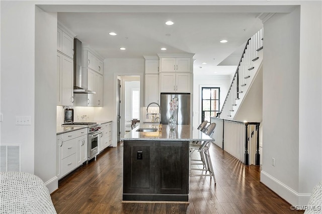 kitchen with an island with sink, wall chimney exhaust hood, appliances with stainless steel finishes, white cabinetry, and a sink