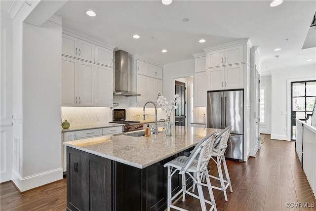 kitchen with high end refrigerator, white cabinets, an island with sink, wall chimney range hood, and a sink