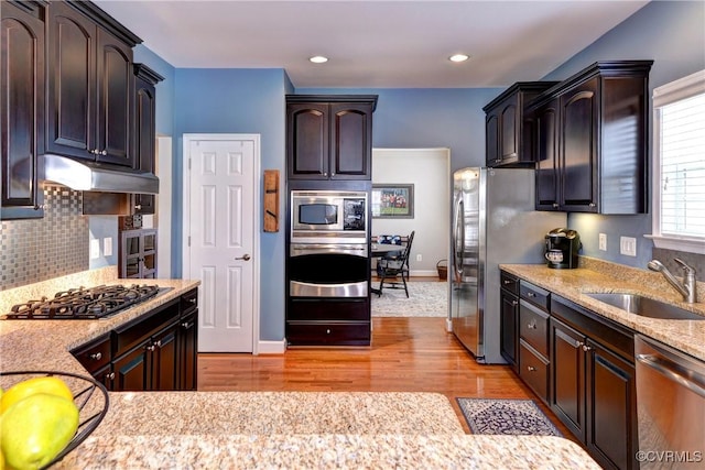 kitchen with dark brown cabinetry, under cabinet range hood, stainless steel appliances, a sink, and backsplash