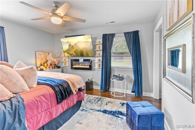 bedroom featuring baseboards, visible vents, a ceiling fan, a glass covered fireplace, and dark wood-style floors