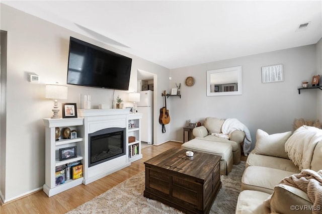 living room featuring light wood-style flooring, a glass covered fireplace, visible vents, and baseboards