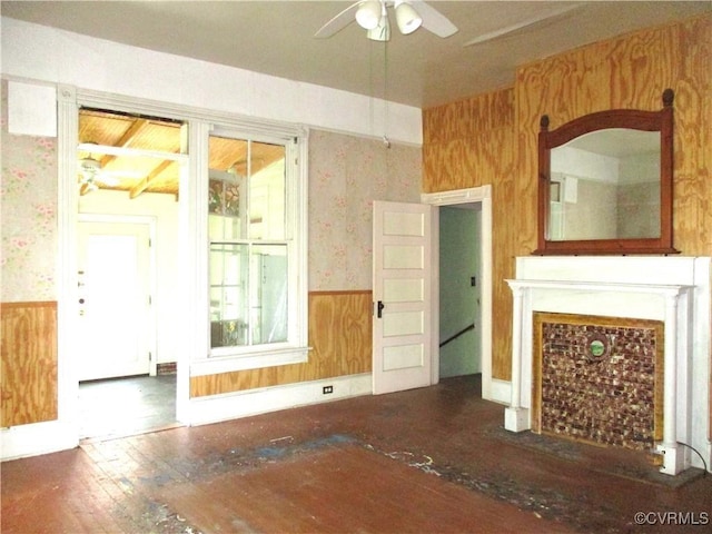 unfurnished living room featuring wooden walls, a ceiling fan, wainscoting, wood-type flooring, and wallpapered walls