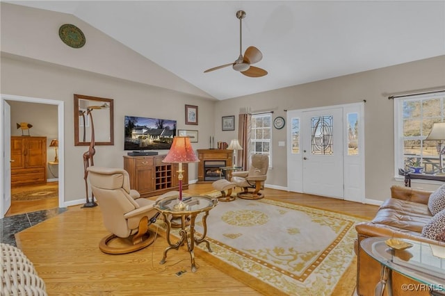 living room featuring high vaulted ceiling, wood finished floors, a ceiling fan, baseboards, and a lit fireplace